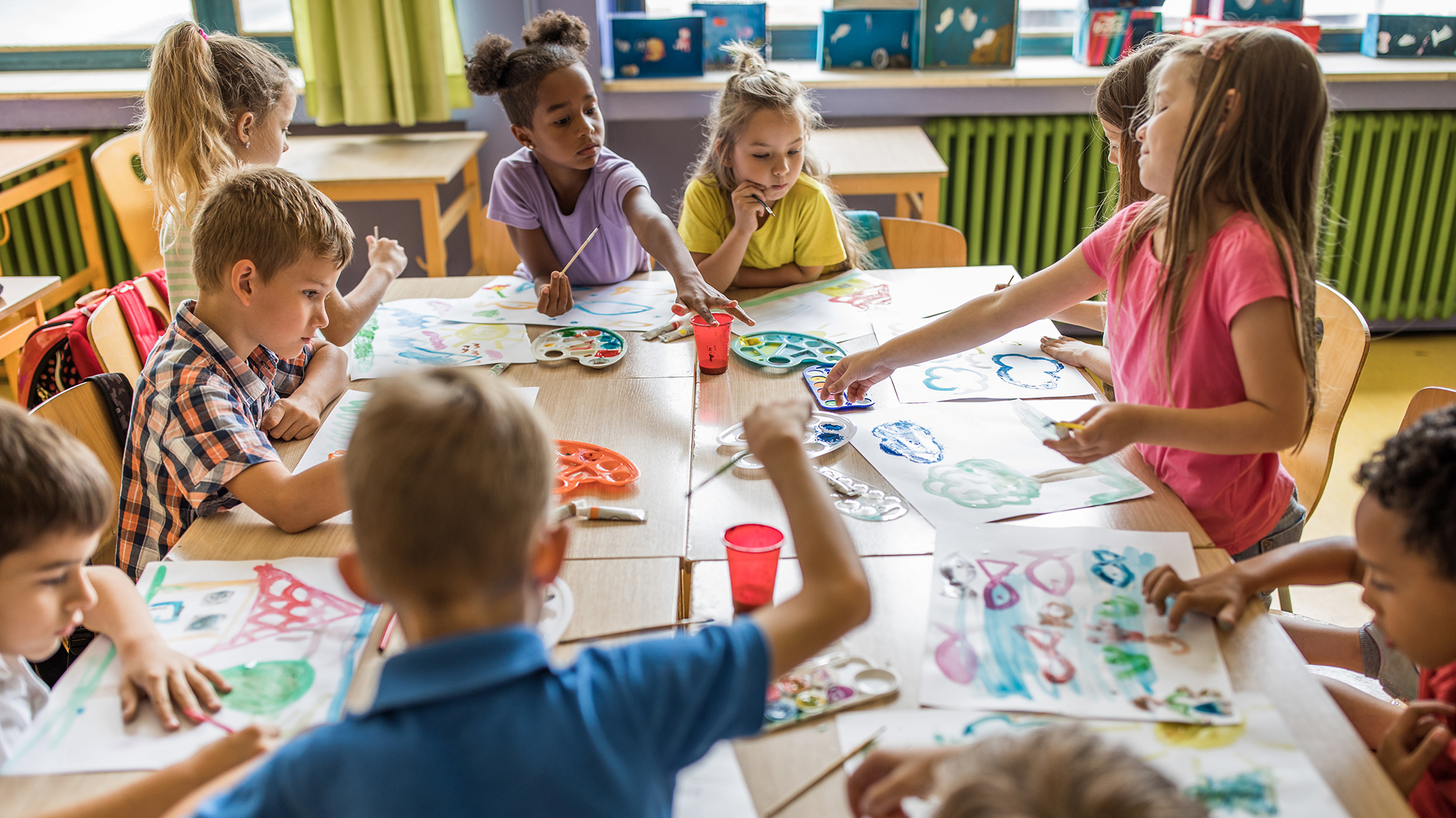  A group of students are sitting around a table in a classroom, painting and collaborating on an art project.