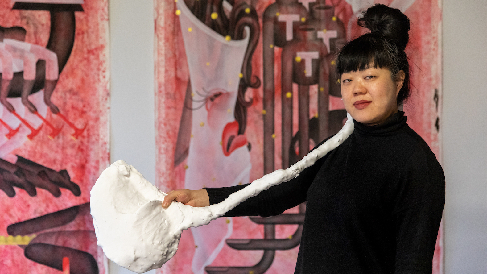 Artist Jen Liu stands in front of a pink and white background holding a long white object.